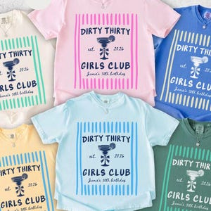 May include: A collection of six t-shirts in various colors, including ivory, pink, blue, and yellow. Each shirt features the text "DIRTY THIRTY GIRLS CLUB" with a cocktail graphic, est. 2026, and "Jamie's 30th birthday".