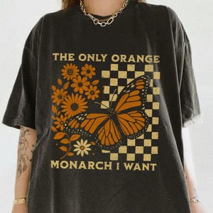 May include: Black t-shirt with the text "THE ONLY ORANGE MONARCH I WANT". The design features an orange monarch butterfly, orange flowers, and a checkered pattern. The shirt is a casual, oversized fit.