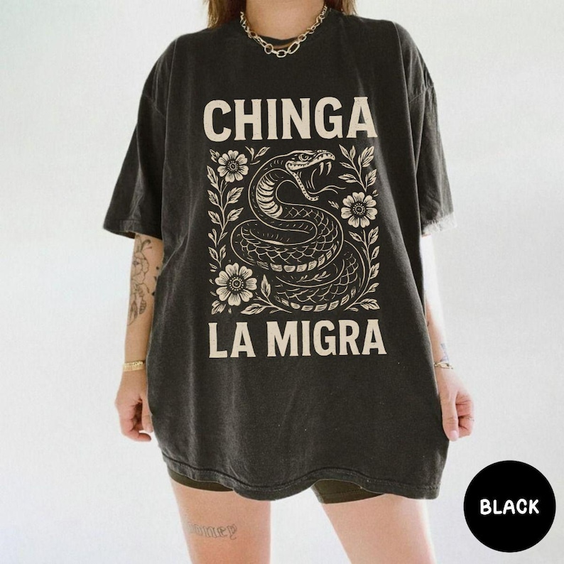 Puede incluir: Camiseta negra con la frase "CHINGA LA MIGRA" en letras blancas. El dise&ntilde;o incluye una detallada ilustraci&oacute;n de una serpiente enmarcada por flores y hojas. La camiseta tiene un aspecto negro descolorido.