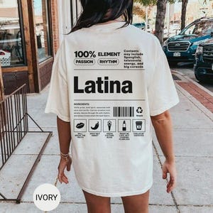 Może przedstawiać: Kremowa koszulka z napisem "Latina" pogrubioną czarną czcionką. Koszulka zawiera tekst "100% ELEMENT" i "PASSION RHYTHM". Dodatkowy tekst to "Contents may include Spanglish, telenovela energy, and big coraz&oacute;n."