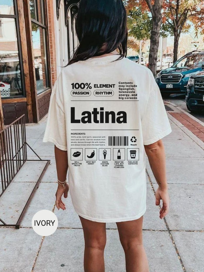 Może przedstawiać: Kremowa koszulka z napisem "Latina" pogrubioną czarną czcionką. Koszulka zawiera tekst "100% ELEMENT" i "PASSION RHYTHM". Dodatkowy tekst to "Contents may include Spanglish, telenovela energy, and big coraz&oacute;n."