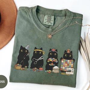 May include: A moss green t-shirt featuring a graphic of four black cats engaged in sewing activities. The cats are depicted with sewing tools including a thimble, scissors, a pincushion, and a quilt. The shirt has a Comfort Colors brand label.