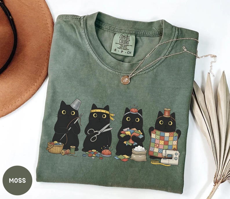 May include: A moss green t-shirt featuring a graphic of four black cats engaged in sewing activities. The cats are depicted with sewing tools including a thimble, scissors, a pincushion, and a quilt. The shirt has a Comfort Colors brand label.