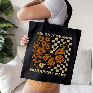 May include: Black tote bag featuring a monarch butterfly with orange and black wings, surrounded by orange flowers and a checkered pattern. Text reads "THE ONLY ORANGE MONARCH I WANT". The bag has black handles.