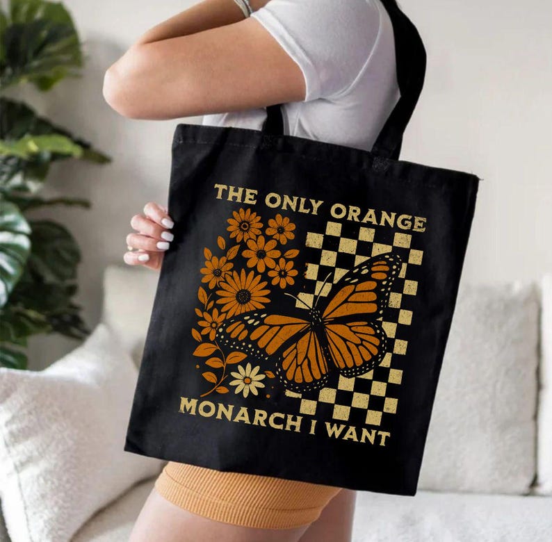 May include: Black tote bag featuring a monarch butterfly with orange and black wings, surrounded by orange flowers and a checkered pattern. Text reads "THE ONLY ORANGE MONARCH I WANT". The bag has black handles.