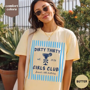 May include: Butter-colored t-shirt with the text "DIRTY THIRTY GIRLS CLUB" in navy blue, with a cocktail graphic. The shirt also includes the text "est. 2026" and "Jamie's 30th birthday" in a script font. The design is framed by blue vertical lines.