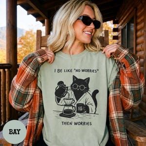 May include: A sage green crewneck sweatshirt featuring a black graphic of a cat wearing round glasses and a robe, pouring coffee into a dripper. The text reads I BE LIKE "NO WORRIES" above the cat and THEN WORRIES below the cat.