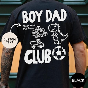 May include: Black t-shirt with white graphic text and illustrations. The text reads "BOY DAD CLUB" with a dinosaur, soccer ball, toy truck, and building blocks. The shirt also has the text "Oliver runs this house".