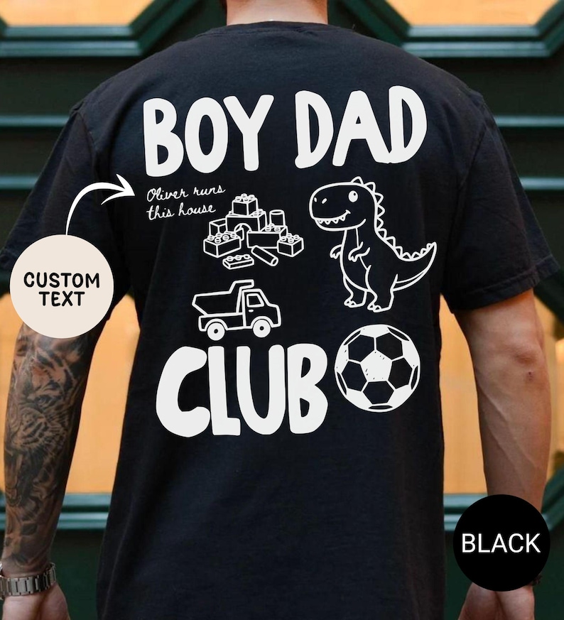 May include: Black t-shirt with white graphic text and illustrations. The text reads "BOY DAD CLUB" with a dinosaur, soccer ball, toy truck, and building blocks. The shirt also has the text "Oliver runs this house".