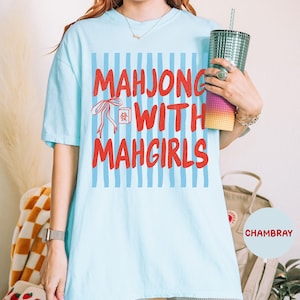 Peut inclure: Un t-shirt bleu clair &agrave; manches courtes avec un fond &agrave; rayures bleues verticales, le texte rouge "MAHJONG WITH MAHGIRLS" et une petite illustration d'un ruban rouge et d'une tuile de mahjong avec un caract&egrave;re chinois imprim&eacute; devant.