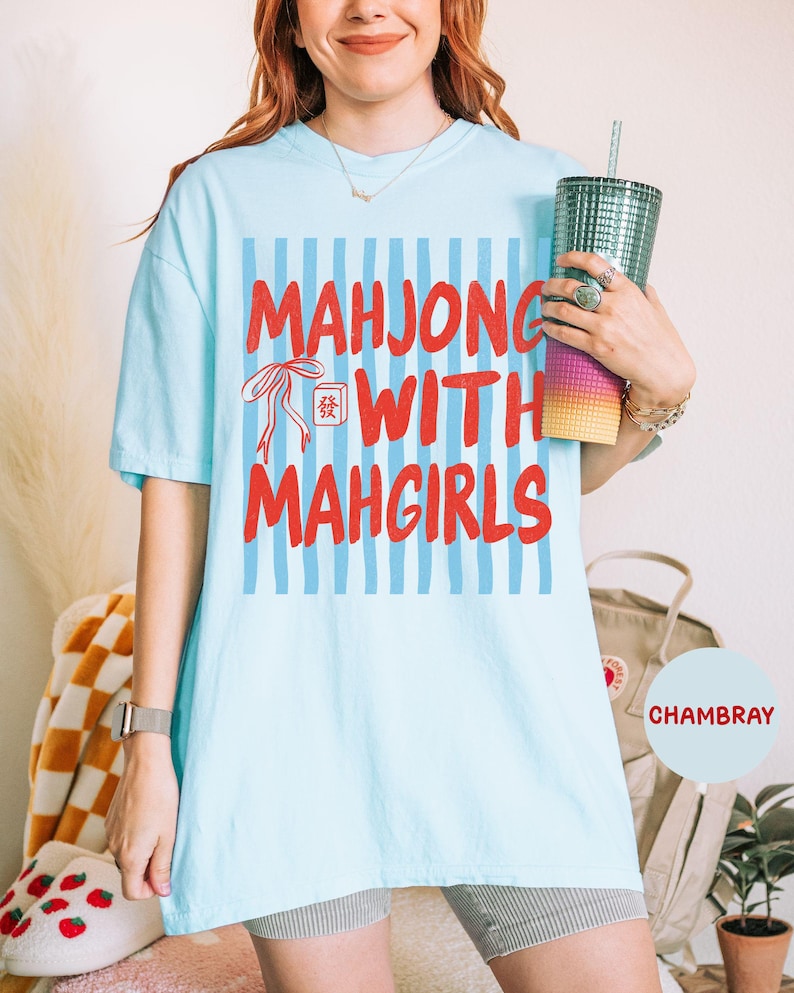 Peut inclure: Un t-shirt bleu clair &agrave; manches courtes avec un fond &agrave; rayures bleues verticales, le texte rouge "MAHJONG WITH MAHGIRLS" et une petite illustration d'un ruban rouge et d'une tuile de mahjong avec un caract&egrave;re chinois imprim&eacute; devant.