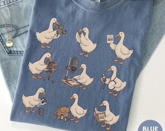 May include: Blue jean Comfort Colors t-shirt featuring a repeating pattern of cartoon geese with cameras and photography equipment. The geese are in various poses, holding cameras, and other media tools. The shirt has a round neck.