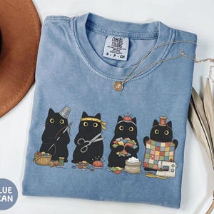 May include: A blue pigment-dyed t-shirt featuring a graphic of four black cats engaged in sewing activities, including holding a needle, scissors, fabric scraps, and a quilt. The shirt has a Comfort Colors label and a crew neckline.