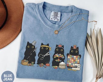 Könnte beinhalten: Ein blaues, pigmentgefärbtes T-Shirt mit einer Grafik von vier schwarzen Katzen, die nähen, darunter eine Nadel, eine Schere, Stoffreste und ein Quilt. Das T-Shirt hat ein Comfort Colors-Etikett und einen Rundhalsausschnitt.