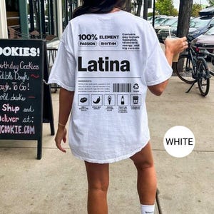 Może przedstawiać: Biała koszulka z napisem "Latina" dużymi czarnymi literami. Tył koszulki zawiera tekst "100% ELEMENT" i "PASSION RHYTHM". Dodatkowy tekst i grafika są wydrukowane poniżej.