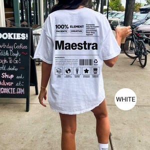 May include: A white t-shirt featuring black text and graphic designs on the back. The print includes the word Maestra, a barcode, and various icons with labels such as Libro Lover, World Connector, Passion Igniter, Creadora de Genios, and Fueled by Cafecito.