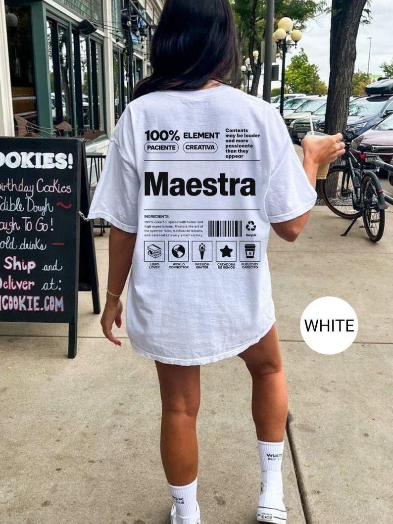 May include: A white t-shirt featuring black text and graphic designs on the back. The print includes the word Maestra, a barcode, and various icons with labels such as Libro Lover, World Connector, Passion Igniter, Creadora de Genios, and Fueled by Cafecito.
