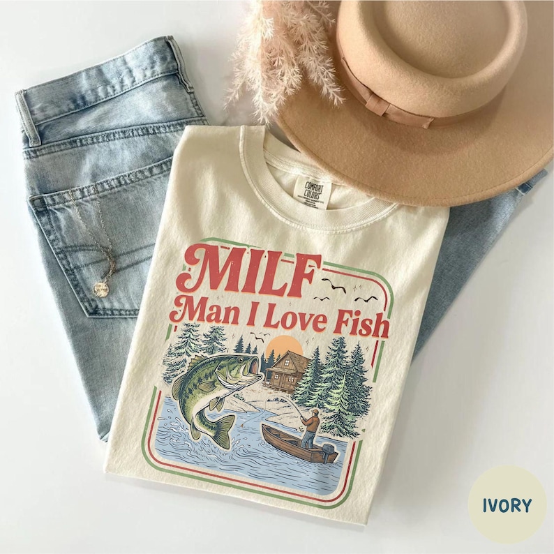 May include: Ivory-colored t-shirt with the text "MILF Man I Love Fish" in red and green. The graphic depicts a fishing scene with a large fish jumping out of the water. Light blue jeans and a tan hat are also in the image.