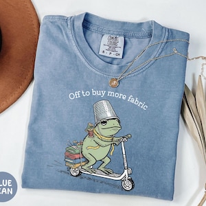 May include: Blue jean Comfort Colors t-shirt with a frog riding a scooter, wearing a thimble hat and carrying spools of thread and fabric. Text reads "Off to buy more fabric."