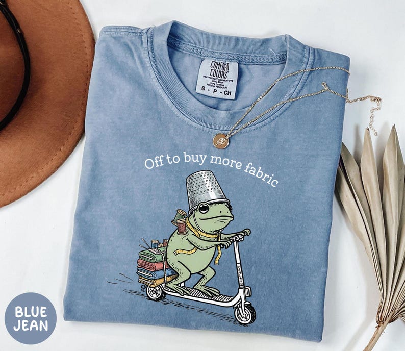 May include: Blue jean Comfort Colors t-shirt with a frog riding a scooter, wearing a thimble hat and carrying spools of thread and fabric. Text reads "Off to buy more fabric."