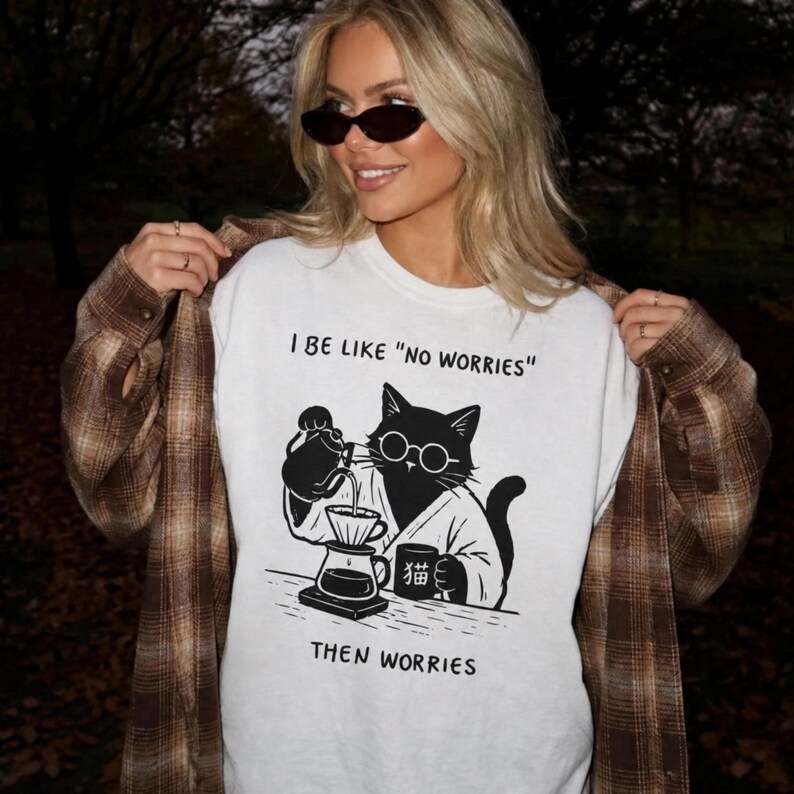 May include: A white t-shirt featuring a black graphic of a cat wearing round glasses and a robe, pouring coffee into a dripper. The text reads I BE LIKE "NO WORRIES" above the cat and THEN WORRIES below. The cat holds a mug with a Japanese character.