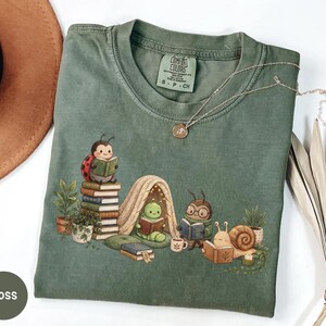 May include: A moss green short-sleeve t-shirt featuring a graphic of a ladybug, a caterpillar in a tent, an insect with glasses, and a snail, all reading books. The shirt has a Comfort Colors label in the neck and a small gold-toned pendant necklace.