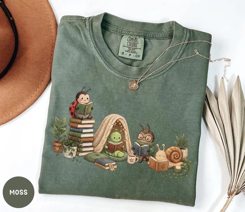 May include: A moss green short-sleeve t-shirt featuring a graphic of a ladybug, a caterpillar in a tent, an insect with glasses, and a snail, all reading books. The shirt has a Comfort Colors label in the neck and a small gold-toned pendant necklace.