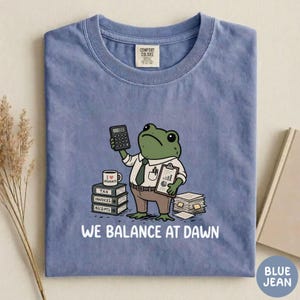 K&ouml;nnte beinhalten: Ein blaues T-Shirt mit einer Grafik eines gr&uuml;nen Frosches in wei&szlig;em Hemd und Krawatte, der einen Taschenrechner und ein Klemmbrett h&auml;lt. Der Frosch steht neben B&uuml;chern mit der Aufschrift Tax, Invoices, Receipts und dem Text WE BALANCE AT DAWN.