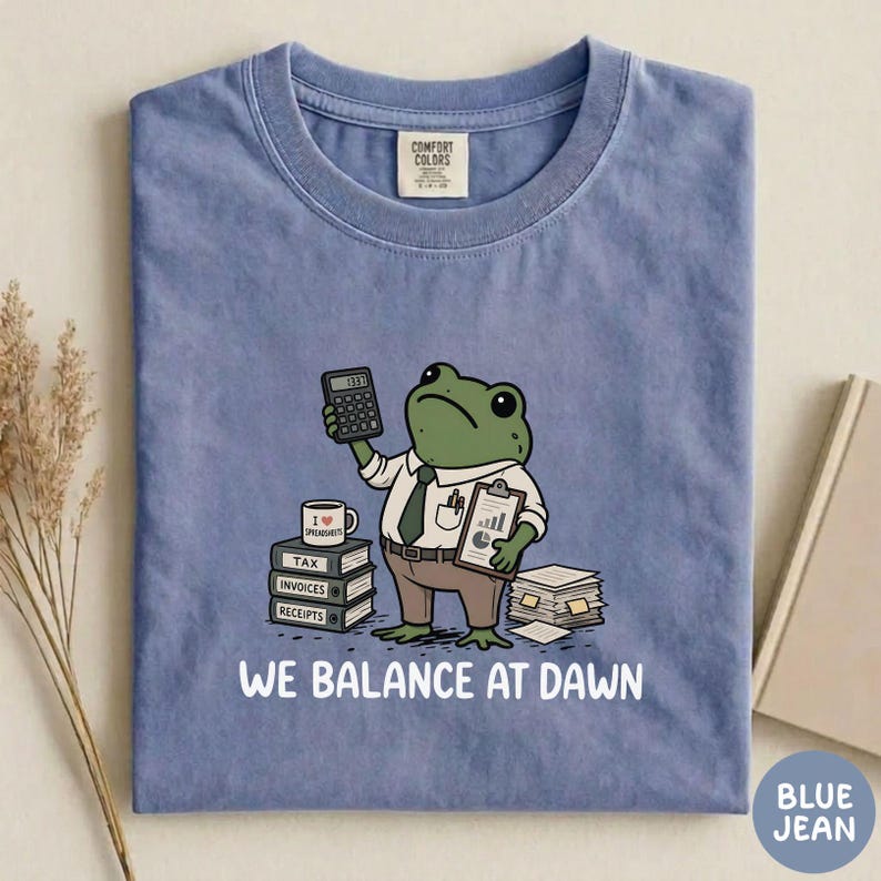 K&ouml;nnte beinhalten: Ein blaues T-Shirt mit einer Grafik eines gr&uuml;nen Frosches in wei&szlig;em Hemd und Krawatte, der einen Taschenrechner und ein Klemmbrett h&auml;lt. Der Frosch steht neben B&uuml;chern mit der Aufschrift Tax, Invoices, Receipts und dem Text WE BALANCE AT DAWN.