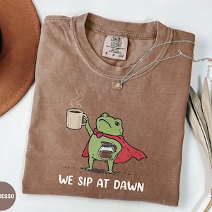 May include: A brown t-shirt featuring a graphic of a green frog wearing a red cape, holding a coffee mug and a coffee pot. The text "WE SIP AT DAWN" is printed in white below the frog. The shirt has a Comfort Colors label at the inner neckline.