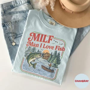 May include: A light blue t-shirt with the text "MILF Man I Love Fish" in red and green. The graphic features a fishing scene with a large fish, a fisherman in a boat, and a cabin in the woods. A tan hat and blue jeans are also in the image.
