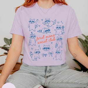 K&ouml;nnte beinhalten: Helllila T-Shirt mit blauen Cartoon-Waschb&auml;ren in verschiedenen Posen. Der Text "feral moms social club" ist in Rot und Blau gedruckt. Das T-Shirt besteht aus einem weichen, bequemen Material.