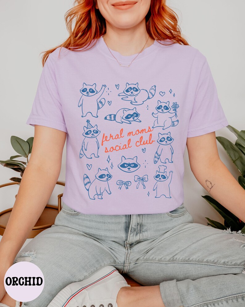 K&ouml;nnte beinhalten: Helllila T-Shirt mit blauen Cartoon-Waschb&auml;ren in verschiedenen Posen. Der Text "feral moms social club" ist in Rot und Blau gedruckt. Das T-Shirt besteht aus einem weichen, bequemen Material.