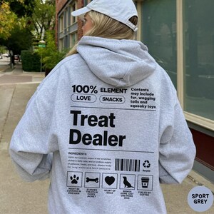 May include: Sport grey hoodie with the text "Treat Dealer" in black. The back of the hoodie features a list of ingredients, including dog hair, treats, and love. The hoodie also has a graphic of a dog and a coffee cup.