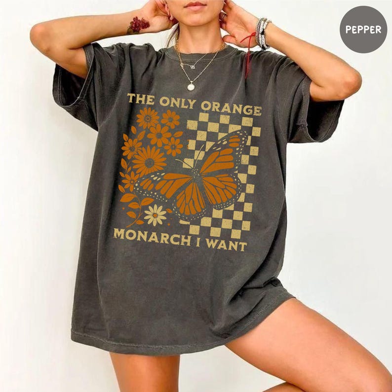 May include: Dark gray t-shirt featuring a monarch butterfly graphic with the text "THE ONLY ORANGE MONARCH I WANT". The design includes orange flowers and a checkered background, creating a vintage aesthetic.