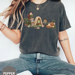 May include: A charcoal gray short-sleeve t-shirt featuring a graphic illustration of a ladybug, a caterpillar in a tent, a beetle with glasses, and a snail, all reading books among potted plants and stacks of books on a light-colored background.