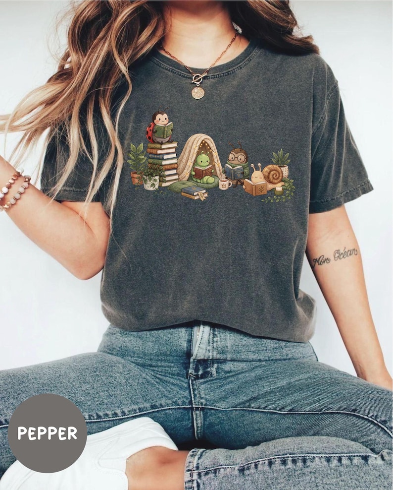May include: A charcoal gray short-sleeve t-shirt featuring a graphic illustration of a ladybug, a caterpillar in a tent, a beetle with glasses, and a snail, all reading books among potted plants and stacks of books on a light-colored background.
