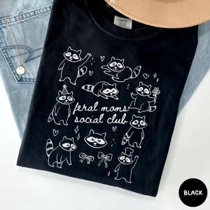 K&ouml;nnte beinhalten: Schwarzes T-Shirt mit einer wei&szlig;en Grafik von Waschb&auml;ren in verschiedenen Posen und dem Text "feral moms social club". Das T-Shirt ist aus weichem Material und wird mit einem Hut und Jeans pr&auml;sentiert. Das T-Shirt ist schwarz.
