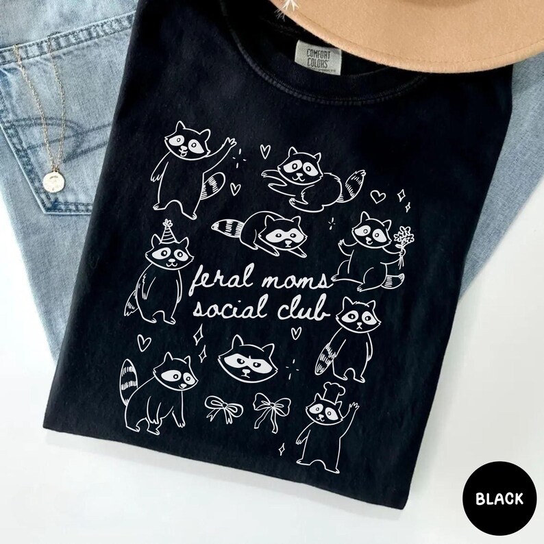 K&ouml;nnte beinhalten: Schwarzes T-Shirt mit einer wei&szlig;en Grafik von Waschb&auml;ren in verschiedenen Posen und dem Text "feral moms social club". Das T-Shirt ist aus weichem Material und wird mit einem Hut und Jeans pr&auml;sentiert. Das T-Shirt ist schwarz.
