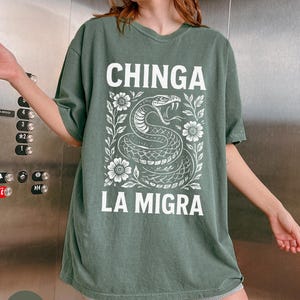 Puede incluir: Camiseta verde musgo con el texto blanco "CHINGA LA MIGRA". Debajo, una ilustraci&oacute;n blanca de una serpiente rodeada de flores y hojas. La camiseta es de corte holgado.