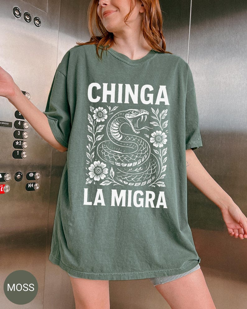 Puede incluir: Camiseta verde musgo con el texto blanco "CHINGA LA MIGRA". Debajo, una ilustraci&oacute;n blanca de una serpiente rodeada de flores y hojas. La camiseta es de corte holgado.