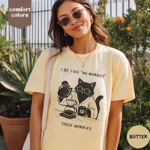 May include: A pale yellow short-sleeve t-shirt featuring a black graphic of a cat wearing glasses and a robe, pouring coffee into a dripper. The text reads I BE LIKE "NO WORRIES" and THEN WORRIES, with a small mug labeled with a Japanese character.