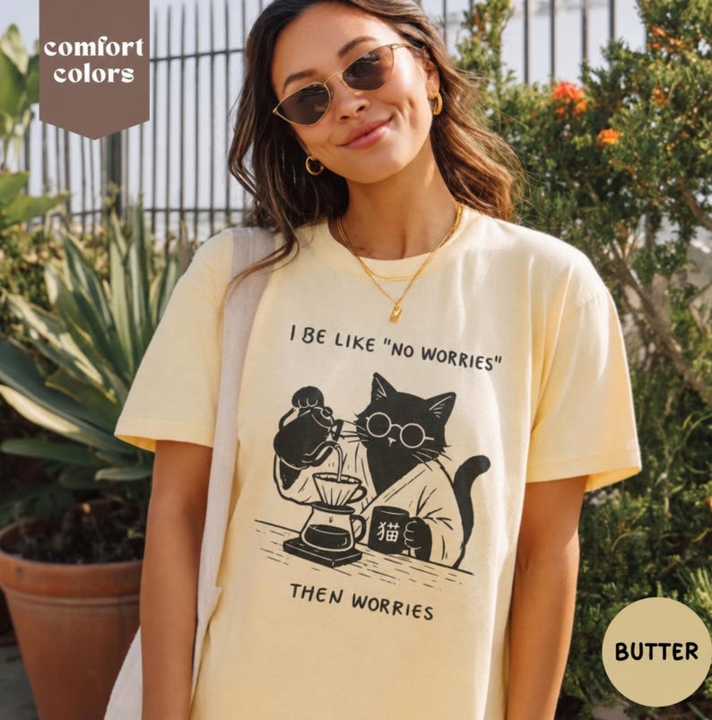 May include: A pale yellow short-sleeve t-shirt featuring a black graphic of a cat wearing glasses and a robe, pouring coffee into a dripper. The text reads I BE LIKE "NO WORRIES" and THEN WORRIES, with a small mug labeled with a Japanese character.