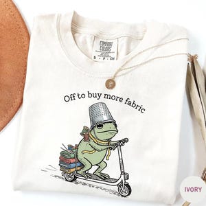 May include: Ivory-colored Comfort Colors t-shirt with the text "Off to buy more fabric." A cartoon frog wearing a thimble hat and carrying spools of thread rides a scooter. A gold necklace is draped on the shirt.