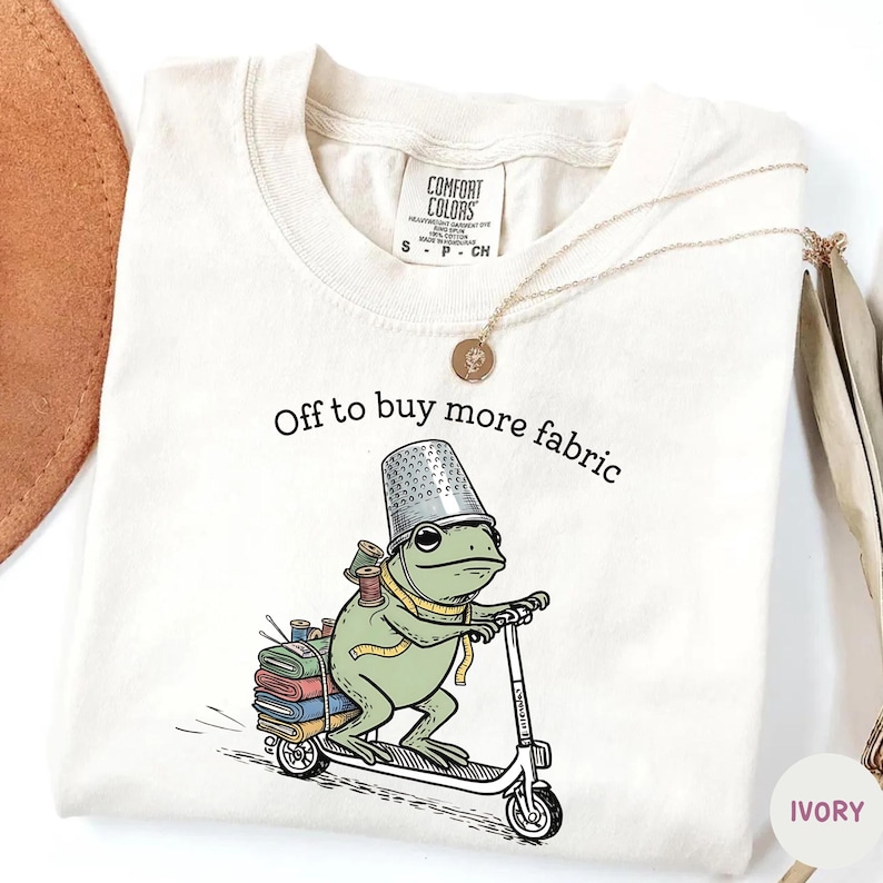 May include: Ivory-colored Comfort Colors t-shirt with the text "Off to buy more fabric." A cartoon frog wearing a thimble hat and carrying spools of thread rides a scooter. A gold necklace is draped on the shirt.