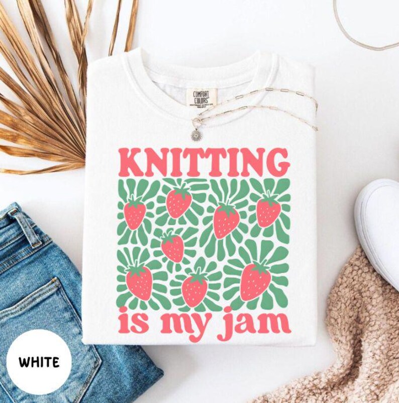 May include: White t-shirt with the text "KNITTING is my jam" in pink. The shirt features a green and white pattern with pink strawberries. A silver necklace and a pair of blue jeans are also visible.