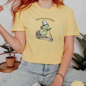 May include: Butter yellow t-shirt featuring a whimsical illustration of a frog riding a scooter, carrying books and a thimble hat. The text "Off to buy more fabric" is printed above the frog. The shirt is a casual, comfortable style.