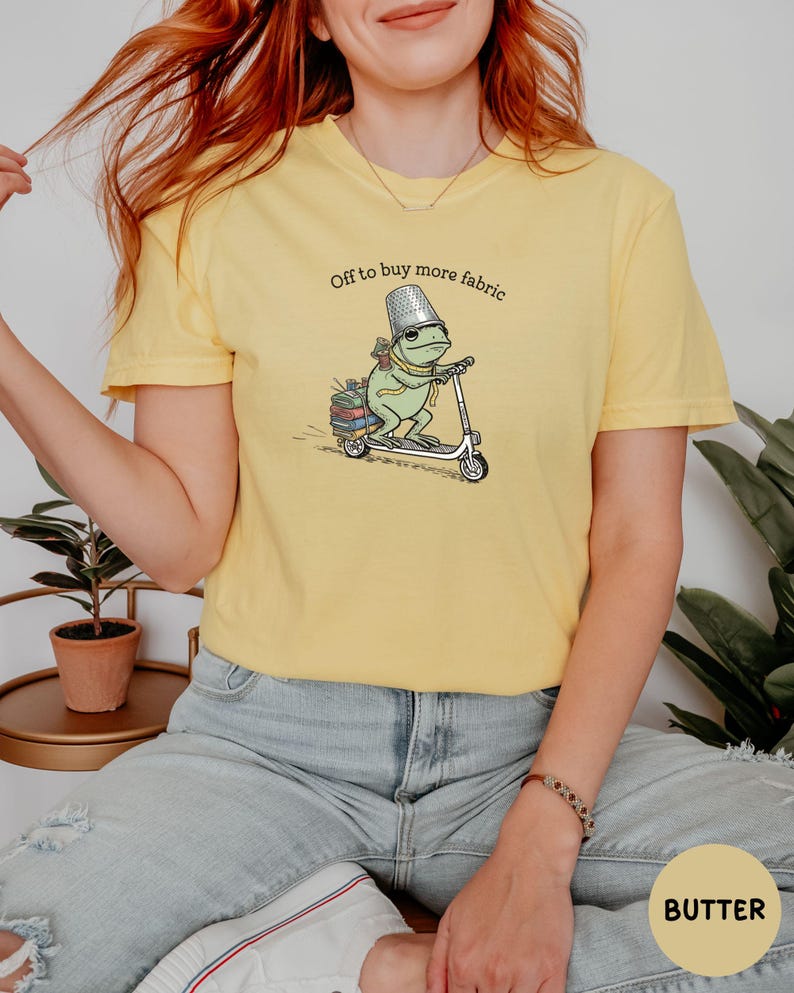 May include: Butter yellow t-shirt featuring a whimsical illustration of a frog riding a scooter, carrying books and a thimble hat. The text "Off to buy more fabric" is printed above the frog. The shirt is a casual, comfortable style.