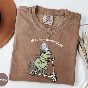 May include: Espresso-colored t-shirt with the text "Off to buy more fabric." The graphic features a frog wearing a thimble hat and riding a scooter, carrying spools of thread and fabric.