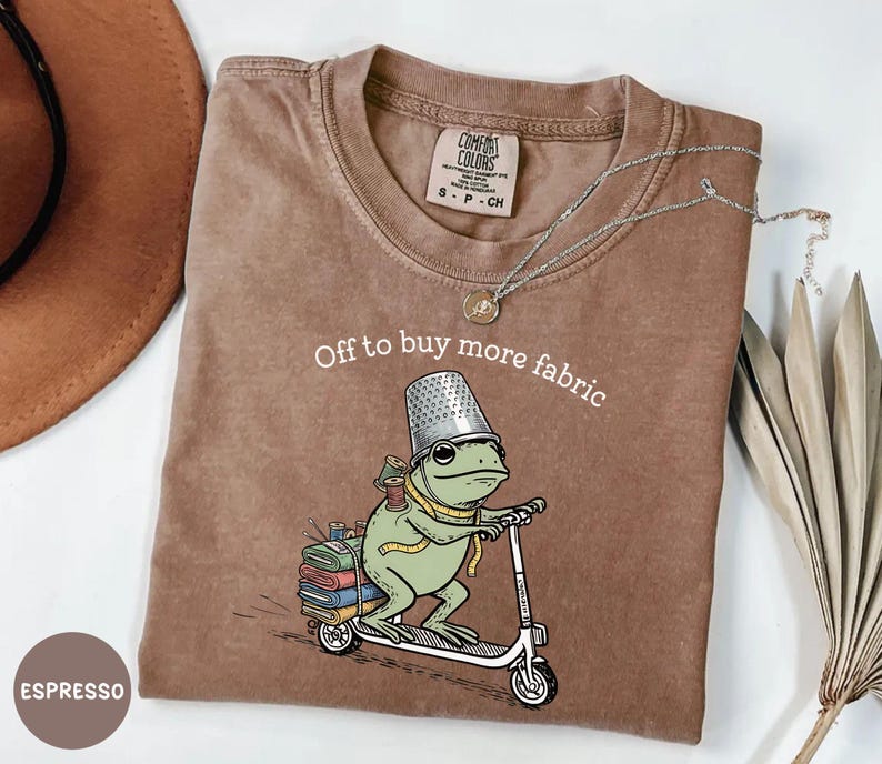 May include: Espresso-colored t-shirt with the text "Off to buy more fabric." The graphic features a frog wearing a thimble hat and riding a scooter, carrying spools of thread and fabric.